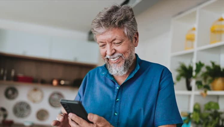 Um senhor de cabelos e barba branca sorrindo e olhando para o celular vestindo uma camisa azul com botões