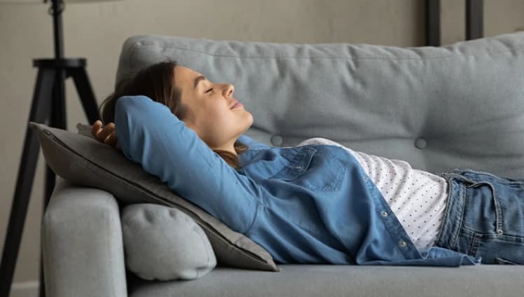 Um mulher branca de cabelos pretos, ela está vestindo uma jaquela de jeans com uma blusa de cor branca e bolinhas pretas por baixo e uma calça jeans, está deitada em um sofá de cor cinza com uma expressão de tranquilidade no rosto