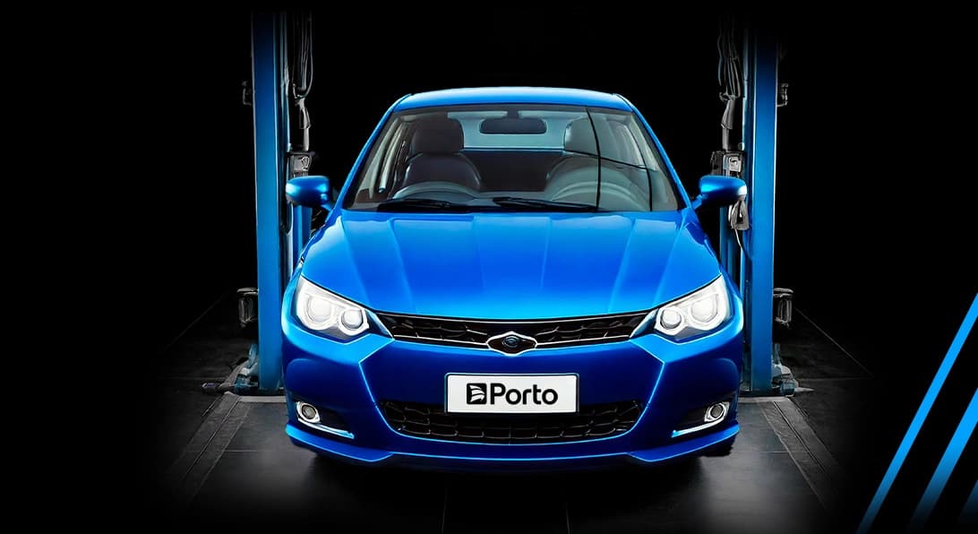 Foto frontal de um carro azul moderno posicionado em um elevador automotivo azul dentro de uma oficina com fundo preto. Os faróis do veículo estão acesos e a placa dianteira exibe o logotipo da Porto.